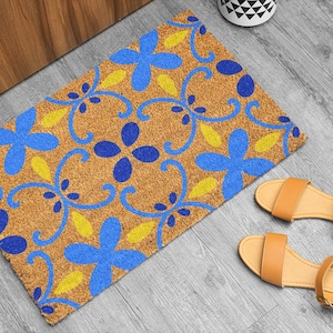 May include: A rectangular doormat with a floral pattern in blue and yellow on a brown background. Two pairs of tan sandals with ankle straps are next to the doormat. The doormat is on a gray wooden floor.
