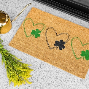 May include: A rectangular coir doormat with three heart designs, each with a shamrock. The hearts are outlined in green and black. A gold watering can and a bundle of yellow flowers are also in the image.
