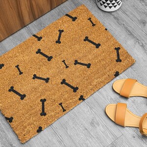 May include: A rectangular doormat with a tan background and black bone pattern. Two pairs of tan sandals with ankle straps are on a gray wooden floor. A white and black patterned container is in the background.