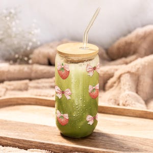 May include: A clear glass can-shaped tumbler filled with a green beverage, featuring a wooden lid and a curved glass straw. The tumbler is decorated with strawberry and bow illustrations. The glass sits on a wooden tray.