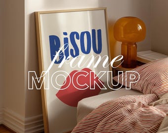 Könnte beinhalten: Gerahmter Kunstdruck-Mockup mit dem Wort "Bisou" in Blau, einer roten Lippe und dem Wort "frame MOCKUP" in Weiß. Der Rahmen lehnt an einer Wand, mit einem Nachttisch und einer Lampe sichtbar.