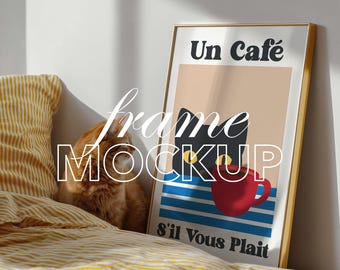 Op de afbeelding: Een ingelijste kunstdruk met de tekst "Un Café" en "s'il Vous Plait" in het zwart. Het kunstwerk toont een zwarte kat, een rode koffiekop en blauw-witte strepen. Een rode kat en een geel gestreept kussen staan op de voorgrond.
