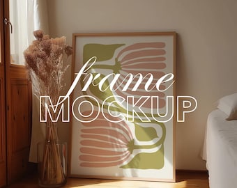 May include: A framed art print featuring a floral design in pink, green, and white. The words "frame MOCKUP" are overlaid on the image. The frame is propped against a wall next to a vase of dried flowers.