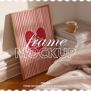 May include: A framed print with a red heart and the words "And so together they built a life they loved" on a red and white striped background. The frame is leaning against a wall. A stack of papers and a mug are in the background. The words "Frame Mockup" are in the image.