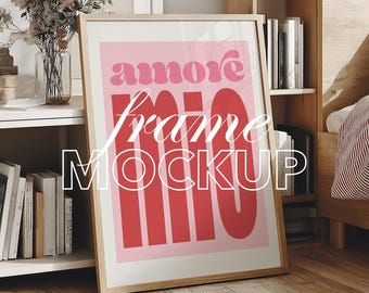 May include: A framed art print with the words "amore mio" in red and pink. The words "frame mockup" are overlaid in white. The frame is light wood, and the background includes a bookshelf and a bed.