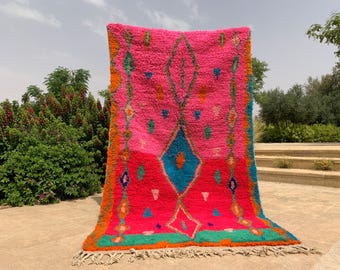 May include: A vibrant, hand-woven rug with a pink central field and a geometric design in turquoise, orange, and green. The rug features a diamond pattern and fringe along the bottom edge. The rug is displayed outdoors.