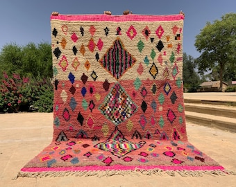 May include: A vibrant, hand-woven rug with a geometric diamond pattern. The rug features a color palette of pink, cream, and various jewel tones. The design includes diamond shapes and a fringed edge, showcasing a traditional Moroccan style.