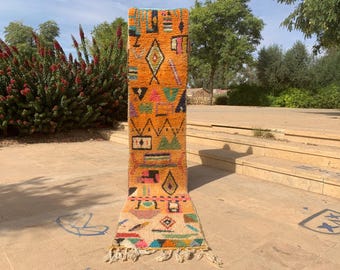 May include: A vibrant, rectangular Moroccan rug with a warm orange background and colorful geometric patterns. The design includes diamonds, triangles, and other shapes in shades of green, pink, and black. The rug has a fringed edge and is displayed outdoors.