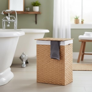 May include: A rectangular woven laundry hamper with a lid and a gray towel hanging over the side. The hamper is light brown with a white interior and is in a bathroom setting.