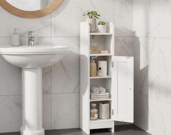 May include: A white, narrow bathroom cabinet with an open door, revealing shelves stocked with toiletries, towels, and decorative items. The cabinet is positioned next to a white pedestal sink and a round mirror. The bathroom has white marble-look tile.