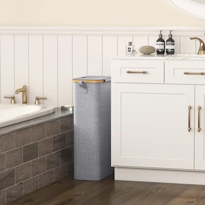 May include: A tall, rectangular gray laundry hamper with a wooden rim and handle. The hamper is made of a textured fabric and is placed next to a white cabinet in a bathroom setting. The bathroom has a bathtub with gold fixtures.