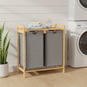 May include: A bamboo laundry hamper with two gray fabric compartments. The hamper has a slatted top shelf holding folded towels and a glass jar. The hamper is in a room with a washing machine and a potted plant.