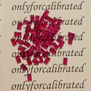 Puede incluir: Un conjunto de pequeñas gemas rectangulares de color rojo intenso, dispersas sobre una superficie blanca. Las gemas están cortadas uniformemente y tienen un aspecto pulido. Las palabras "onlyforcalibrated" están impresas en tinta negra.