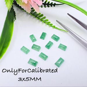 May include: A collection of ten small, rectangular, light green gemstones with a smooth, polished finish. Each stone measures 3x5mm. A pair of silver metal tweezers is positioned to the right of the stones, and green foliage is visible above.