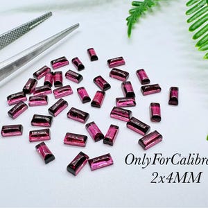 May include: A collection of small, rectangular, deep pink gemstones scattered on a white surface. The stones are uniform in shape and size, with text printed in the corner reading OnlyForCalibrated 2x4MM. Metal tweezers are visible at the top left.