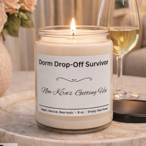 Puede incluir: Una vela de cera de soja natural encendida en un tarro de vidrio con el texto "Dorm Drop-Off Survivor" y "Now Kira's Getting Hot". La vela est&aacute; sobre una bandeja de m&aacute;rmol con una copa de vino y un jarr&oacute;n de rosas rosas.