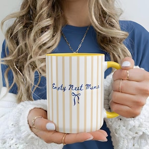 Puede incluir: Una taza de cerámica blanca con rayas beige verticales y un interior y asa amarillos. La taza tiene el texto "Empty Nest Mama" en escritura azul con un lazo azul. La taza es sostenida por una persona que lleva una camisa azul y un cárdigan blanco.