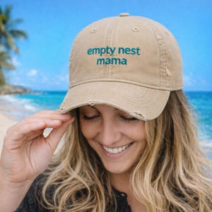 Puede incluir: Gorra de béisbol beige con la inscripción "empty nest mama" bordada en turquesa. La gorra tiene un aspecto desgastado con bordes deshilachados. El fondo muestra una escena de playa con mar y cielo azules.