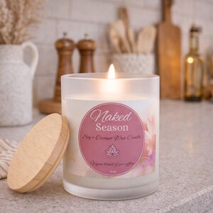 May include: A lit soy and coconut wax candle in a frosted glass jar with a wooden lid. The candle has a pink label with the words "Naked Season" and a floral design. The candle is 11 oz.