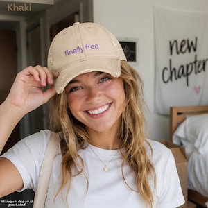 Puede incluir: Una gorra de béisbol color caqui con un acabado desgastado y el texto finally free bordado en morado en el panel frontal. La gorra presenta una visera curva y una corona estructurada, mostrando un diseño casual con bordes deshilachados.