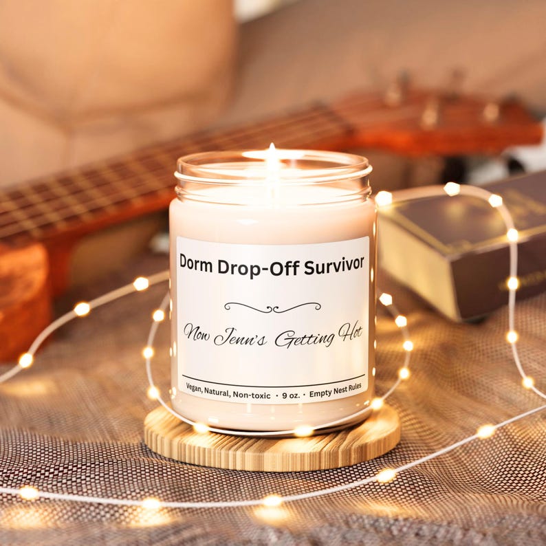 Puede incluir: Una vela encendida en un tarro de cristal con el texto "Dorm Drop-Off Survivor" y "Now Jenn's Getting Hot". La vela est&aacute; sobre un posavasos redondo de madera. Una cadena de luces y un ukelele est&aacute;n en el fondo.