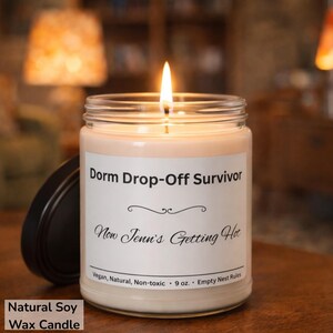 Puede incluir: Una vela de cera de soja encendida en un tarro de vidrio con tapa negra. La etiqueta dice "Dorm Drop-Off Survivor" y "Now Jenn's Getting Hot". La vela es de 255 gramos y es vegana, natural y no t&oacute;xica.