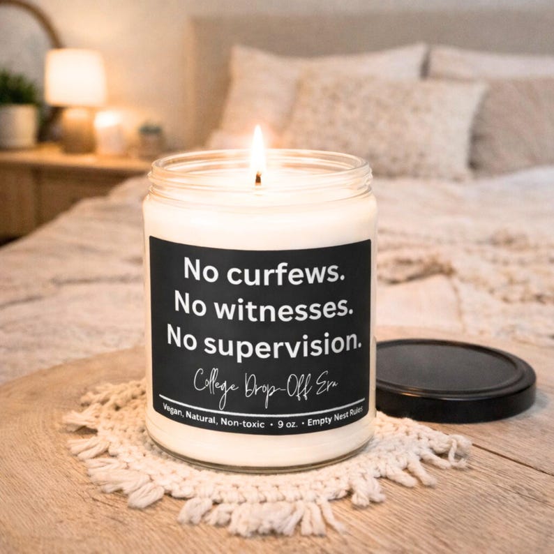 May include: A lit candle in a clear glass jar with a black label. The label reads "No curfews. No witnesses. No supervision." with "College Drop-Off Era" below. The candle is on a white doily.