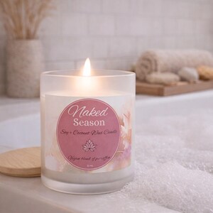 May include: A lit, frosted glass candle labeled "Naked Season" with a pink and floral design. The candle is a soy and coconut wax blend, and the label includes a lotus flower graphic. The candle is 11 oz.