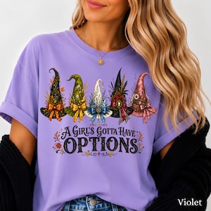 May include: A violet short-sleeved t-shirt featuring a graphic of five distinct witch hats in black, green, white, dark, and pink colors. Below the hats, the text reads A GIRL'S GOTTA HAVE OPTIONS in black lettering with decorative floral accents.