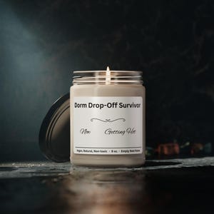 Puede incluir: Una vela encendida en un frasco de vidrio con tapa plateada. La etiqueta dice "Dorm Drop-Off Survivor" con las frases "Now Getting Hot" debajo. La vela es de 255 gramos y es vegana, natural y no t&oacute;xica.