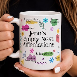 May include: A white ceramic mug featuring the black text Jenn's Empty Nest Affirmations, surrounded by colorful floral illustrations and phrases including I parented hard. I play harder., Zero supervision., Kids grown. Bra optional., and Badass unlocked.