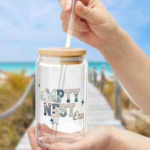 May include: A clear glass tumbler with a bamboo lid and a clear straw. The tumbler features the text "EMPTY NEST Era" in blue and white lettering. The tumbler is held in front of a blurred beach background.