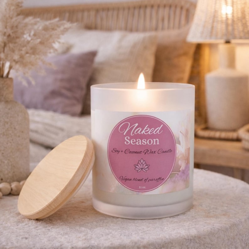 May include: A lit white candle with a wooden lid. The candle has a label that reads "Naked Season Soy + Coconut Wax Candle". The candle is in a frosted glass jar. The candle is 11 oz.