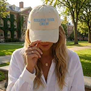 May include: A cream-colored baseball cap with a distressed brim and the text COLLEGE TOUR CREW embroidered in light blue on the front. The cap features a structured crown, eyelets for ventilation, and a button on top, providing a casual look.