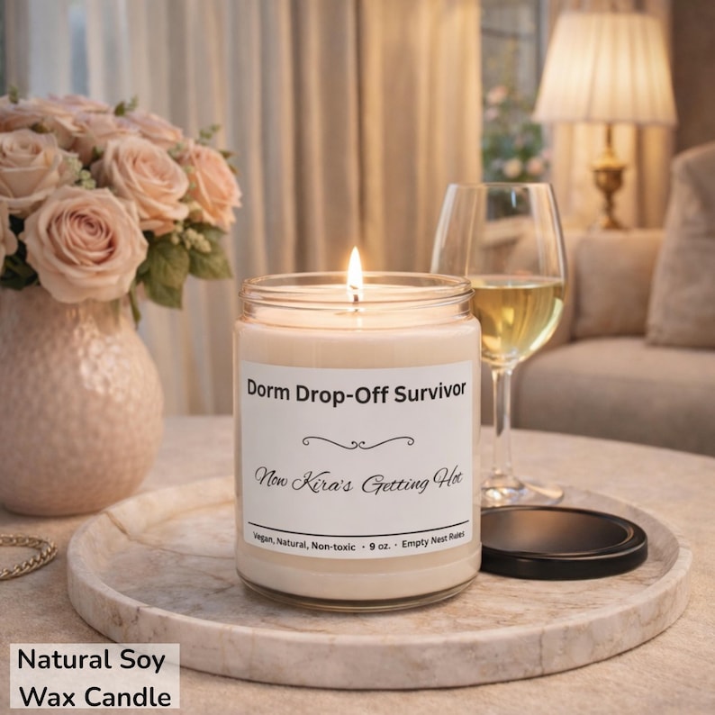 Puede incluir: Una vela de cera de soja natural encendida en un tarro de vidrio con el texto "Dorm Drop-Off Survivor" y "Now Kira's Getting Hot". La vela est&aacute; sobre una bandeja de m&aacute;rmol con una copa de vino y un jarr&oacute;n de rosas rosas.