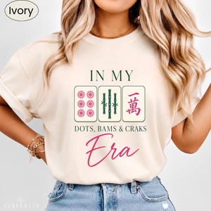 May include: An ivory t-shirt featuring a graphic with three mahjong tiles and the text IN MY DOTS, BAMS & CRAKS Era. The centre tile displays green bamboo stalks, while the flanking tiles show pink circular patterns and a pink Chinese character.