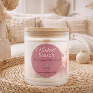May include: A soy and coconut wax candle in a frosted glass jar with a wooden lid. The label reads "Naked Season" in script, with a lotus flower graphic. The candle sits on a woven tray, with a small decorative vase nearby.