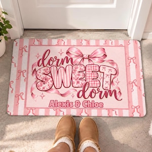 May include: A rectangular doormat featuring a pink and white striped background with a repeating bow pattern. The center displays the text "dorm SWEET dorm" in a stylized font, with the names "Alexis & Chloe" printed below in a dark pink color.