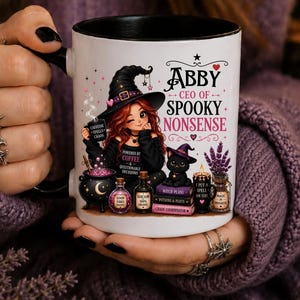 May include: A white ceramic mug with a black handle and interior featuring a graphic of a red-haired witch in a black hat, a black cat, a cauldron, potion bottles, and books. The text reads ABBY CEO OF SPOOKY NONSENSE, CAFFEINE SPELLS CHAOS, and more.
