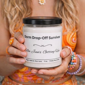 Puede incluir: Vela blanca en un tarro de vidrio con tapa negra. La etiqueta dice "Dorm Drop-Off Survivor" y "Now Jenn's Getting Hot". El tarro se sostiene frente a una persona que usa ropa y joyas de color naranja y amarillo. La vela es de 255 gramos.