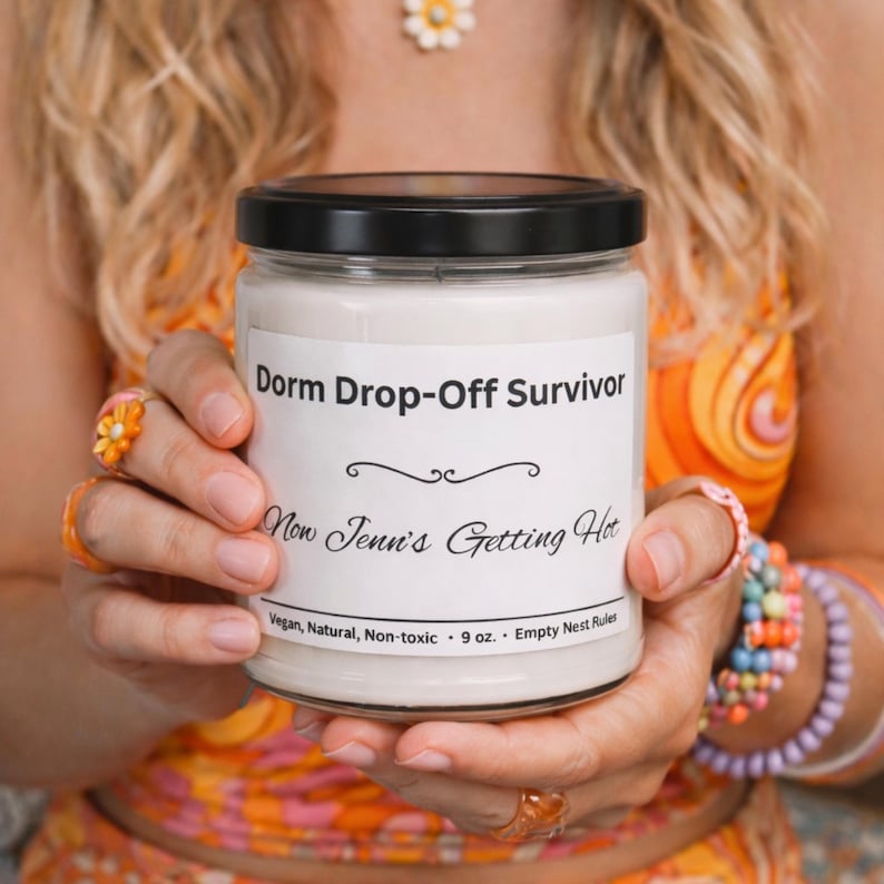 Puede incluir: Vela blanca en un tarro de vidrio con tapa negra. La etiqueta dice "Dorm Drop-Off Survivor" y "Now Jenn's Getting Hot". El tarro se sostiene frente a una persona que usa ropa y joyas de color naranja y amarillo. La vela es de 255 gramos.