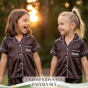 Puede incluir: Dos ni&ntilde;os con conjuntos de pijama de sat&eacute;n marr&oacute;n a juego con ribetes blancos y nombres personalizados bordados en el bolsillo. El texto "CUSTOM KIDS SATIN PAJAMA SET" es visible en la parte inferior de la imagen.