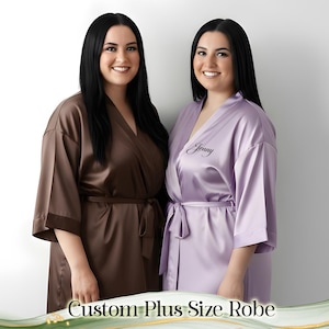 May include: Two women wearing satin robes. One robe is brown, the other is lavender with the name "Jenny" embroidered on the front. The robes have a tie closure and wide sleeves. The text "Custom Plus Size Robe" is at the bottom.
