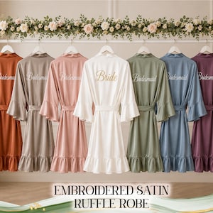 Puede incluir: Una colección de siete batas de satén con dobladillos y mangas con volantes, colgadas en un perchero. Seis batas son de varios colores apagados con la palabra Bridesmaid bordada en la espalda, y una bata blanca tiene la palabra Bride en oro.