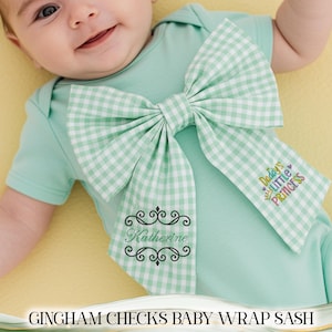 May include: A mint green baby bodysuit with a large gingham bow. The bow is white and light green. The text "Daddy's Little Princess" is embroidered on the bow. The name "Katherine" is embroidered on the bodysuit.