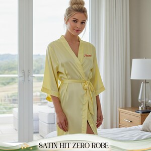 May include: A pale yellow satin robe with a matching tie belt. The robe features a wrap-around style and short sleeves. The name "Naomi" is embroidered in red on the chest. The text "SATIN HIT ZERO ROBE" is at the bottom of the image.