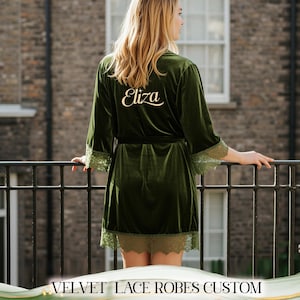 May include: A dark green velvet robe with lace trim, personalized with the name "Eliza" in gold script on the back. The robe has a matching belt and is knee-length. The image is of a person standing on a balcony.