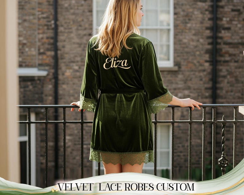May include: A dark green velvet robe with lace trim, personalized with the name "Eliza" in gold script on the back. The robe has a matching belt and is knee-length. The image is of a person standing on a balcony.