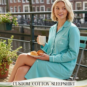 Könnte beinhalten: Hellblaues, maßgeschneidertes Baumwoll-Nachthemd mit weißer Paspelierung und dem Namen "Sia" auf der Tasche. Das Nachthemd mit Knopfleiste wird von einer Person getragen, die draußen mit einer Tasse und einem Teller Gebäck sitzt.