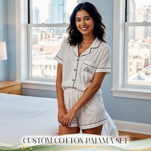 Peut inclure: Ensemble de pyjama en coton blanc avec bordure noire, composé d'un haut à manches courtes et d'un short. Le haut a une poche poitrine avec le nom "Ellie" brodé. Le texte "CUSTOM COTTON PAJAMA SET" est au bas de l'image.